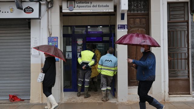 Varias personas hacen cola en una administración de Lotería en Catarroja bajo la lluvia. / MANUEL BRUQUE (EFE)